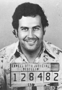 Black-and-white mugshot-style photo of Eric Nicholas, author of Refugium, smiling at the camera while holding a sign displaying identification details.